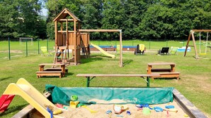 Un parque infantil con una casita de juegos de madera, toboganes, columpios, mesas de picnic y un arenero lleno de juguetes.