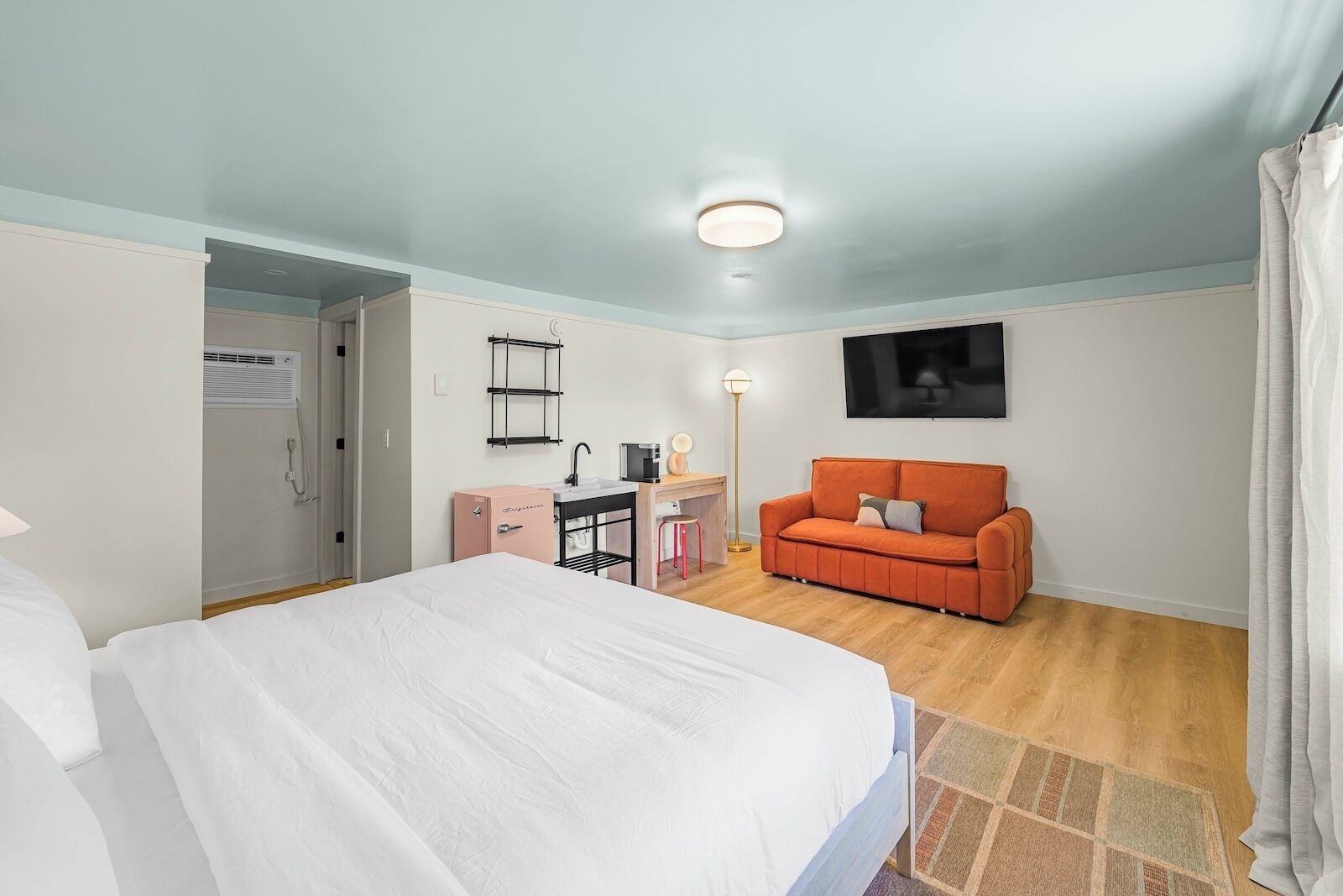 Habitación de hotel con cama, escritorio con silla, sofá, televisor y kitchenette pequeña.