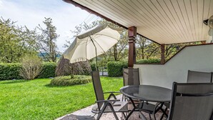 Un patio exterior con una mesa y sillas, un paraguas grande y vistas a un jardín cuidado.
