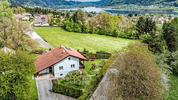 Una casa con techo rojo, rodeada de césped cuidado y árboles, con vistas a un lago y montañas.