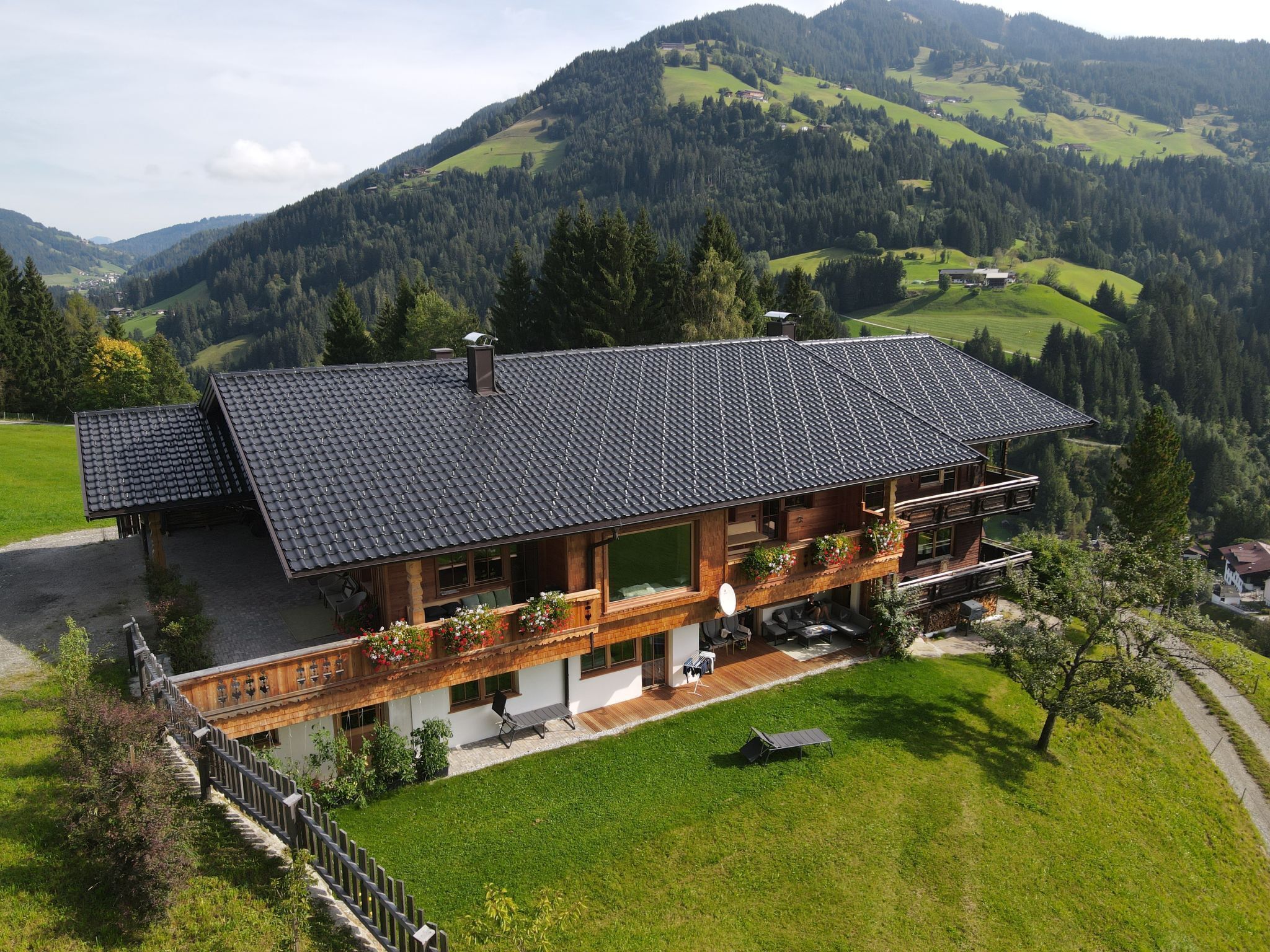 A large, two-story chalet with a sloped roof, surrounded by lush green hills and a forested mountain backdrop.