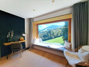 Ein Zimmer mit großem Fenster, Schreibtisch mit Lampe, Stuhl und Sofa mit Blick auf Wald und Berge.