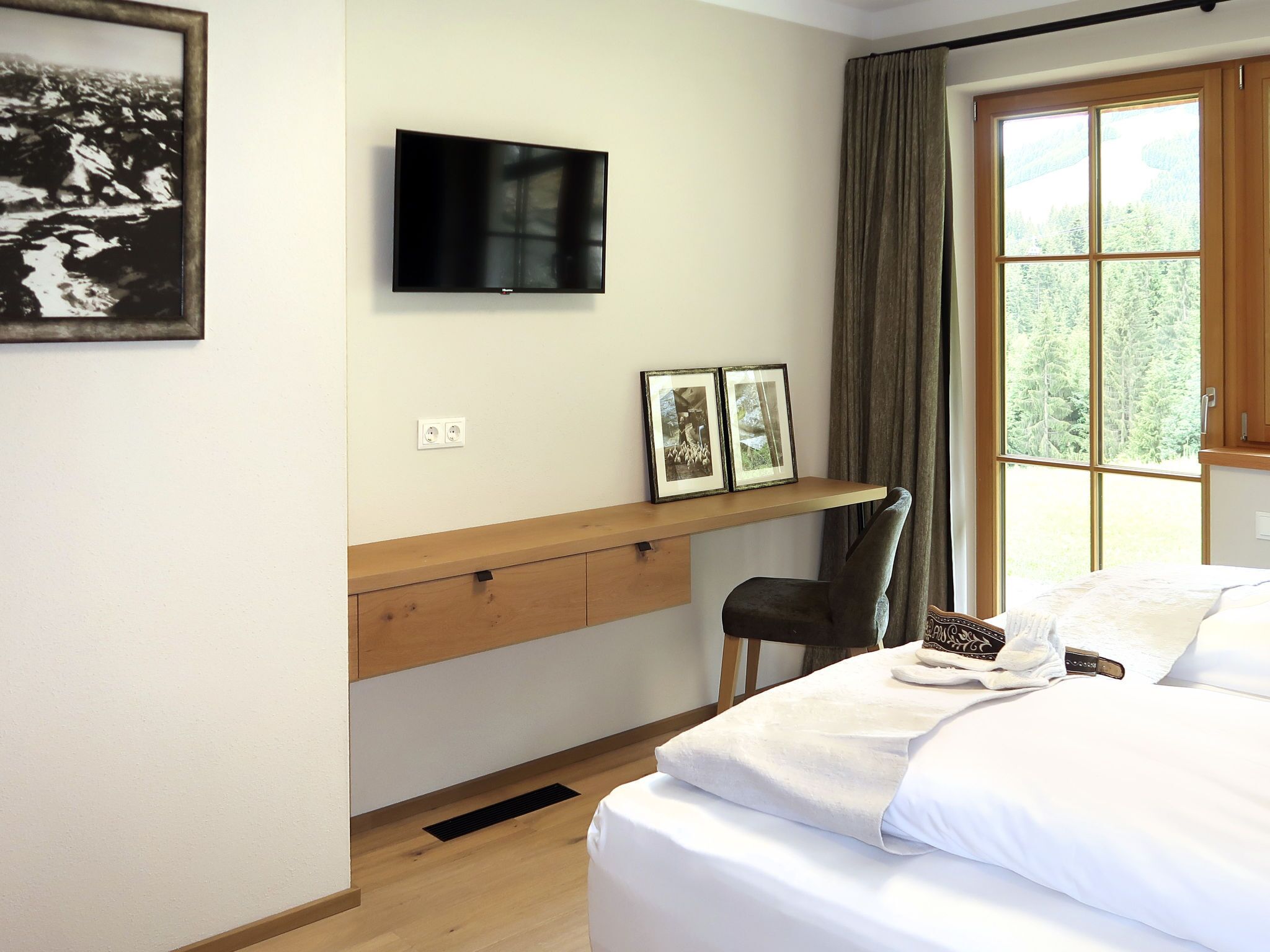 Une chambre à coucher comprenant un lit, un téléviseur fixé au mur, un bureau avec une chaise et une fenêtre avec des rideaux.
