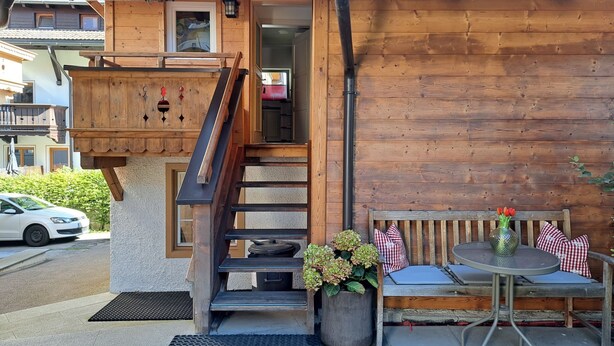 Un edificio de madera con una escalera que conduce a una entrada, un banco con cojines y una mesa pequeña con un jarrón de flores.