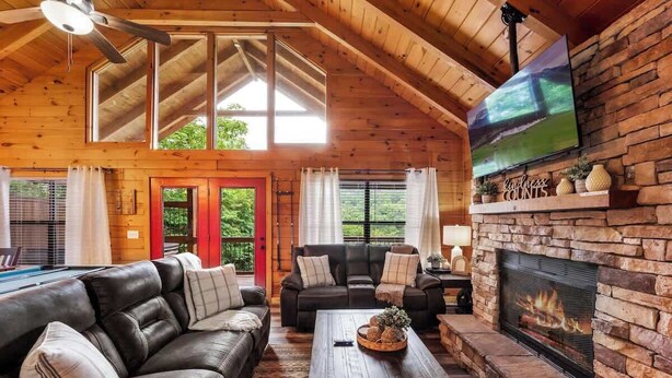 Interior d'una cabana acollidora amb una llar de pedra, sofàs de cuir i un televisor gran penjat a la paret.