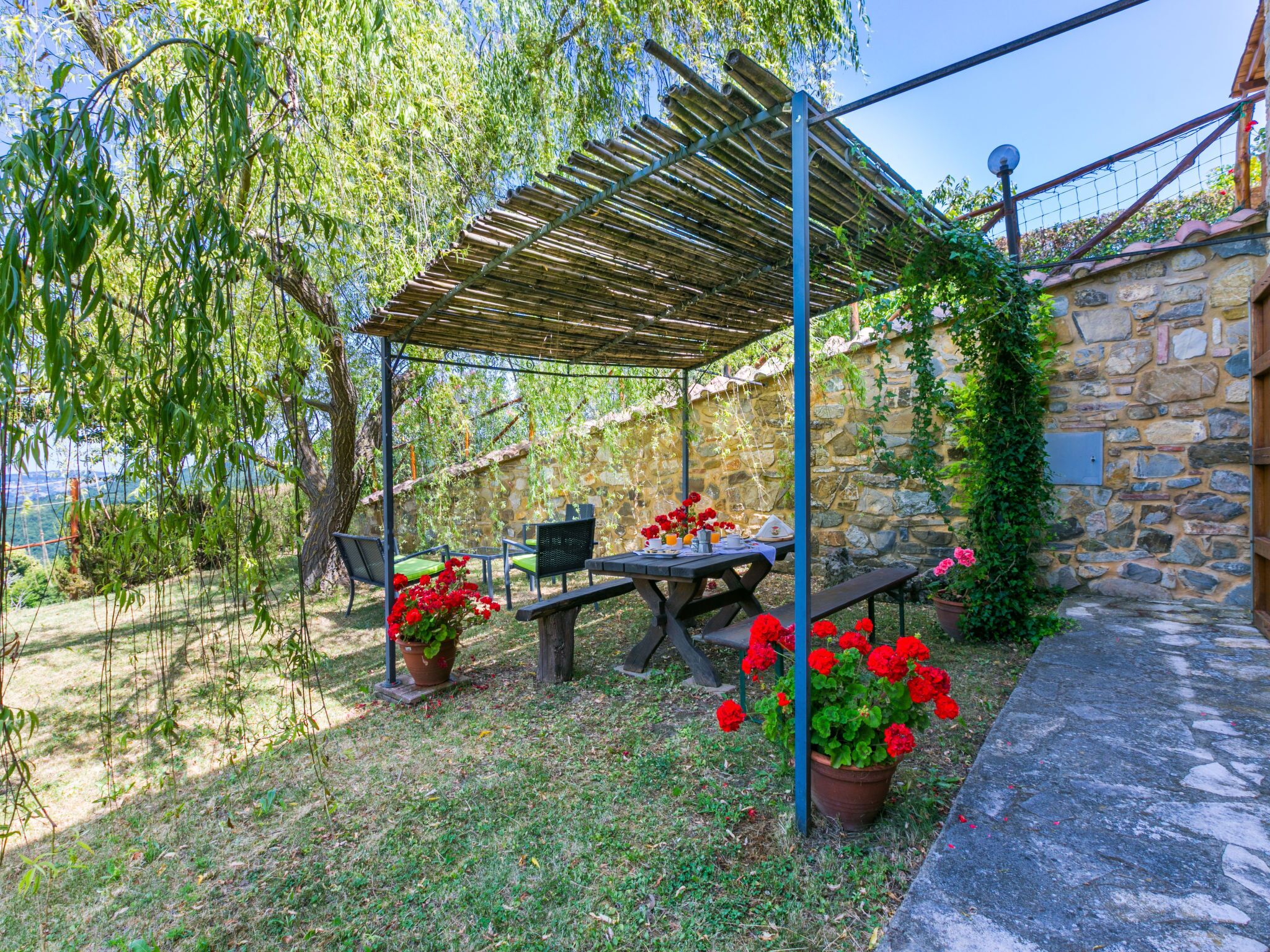Un área de estar al aire libre con techo, un muro de piedra de fondo, una mesa con sillas y plantas en macetas.