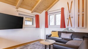 A modern living room with a leather sofa, a small round table, and a TV mounted on the wall. There are windows with red curtains and a wooden ceiling. - für bis zu 8 Personen + Hot Tub by Interhome (Turracher Höhe)