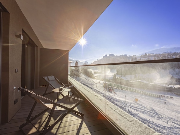 A balcony with sun lounger chairs, a glass railing, and a view of a snowy landscape with buildings and trees.