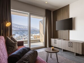A modern hotel room with a large window, a flat-screen TV, a sofa, and a small table.