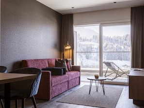 Een moderne woonkamer met een bank, een eettafel en een balkon met uitzicht op besneeuwde bergen.