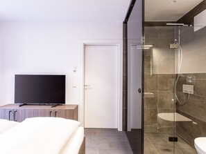 Moderni hotellihuone, jossa on sänky, litteänäyttöinen televisio puukehikossa, lasiseinäinen suihku ja valkoinen ovi.