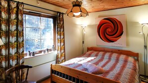 Una cama de madera con colcha estampada, una silla de madera, una ventana con cortinas y una pared con un cuadro abstracto grande.
