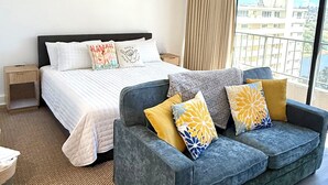 Camera d'albergo con un letto, un divano grigio con cuscini decorati, un comodino in legno e un'ampia finestra con vista sulla città.
