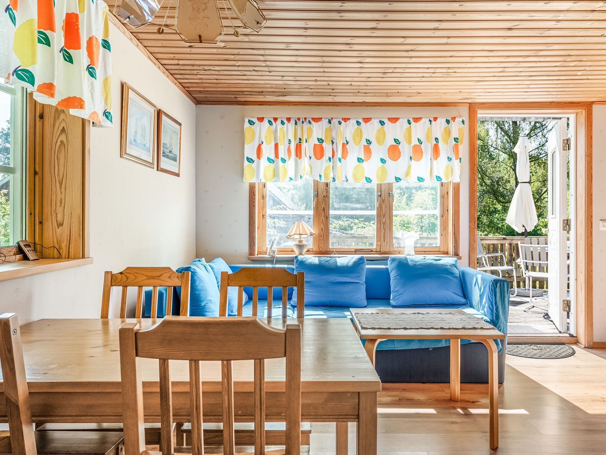 A cozy room with a wooden ceiling, a blue sofa, a wooden dining table, and a window with fruit-patterned curtains.