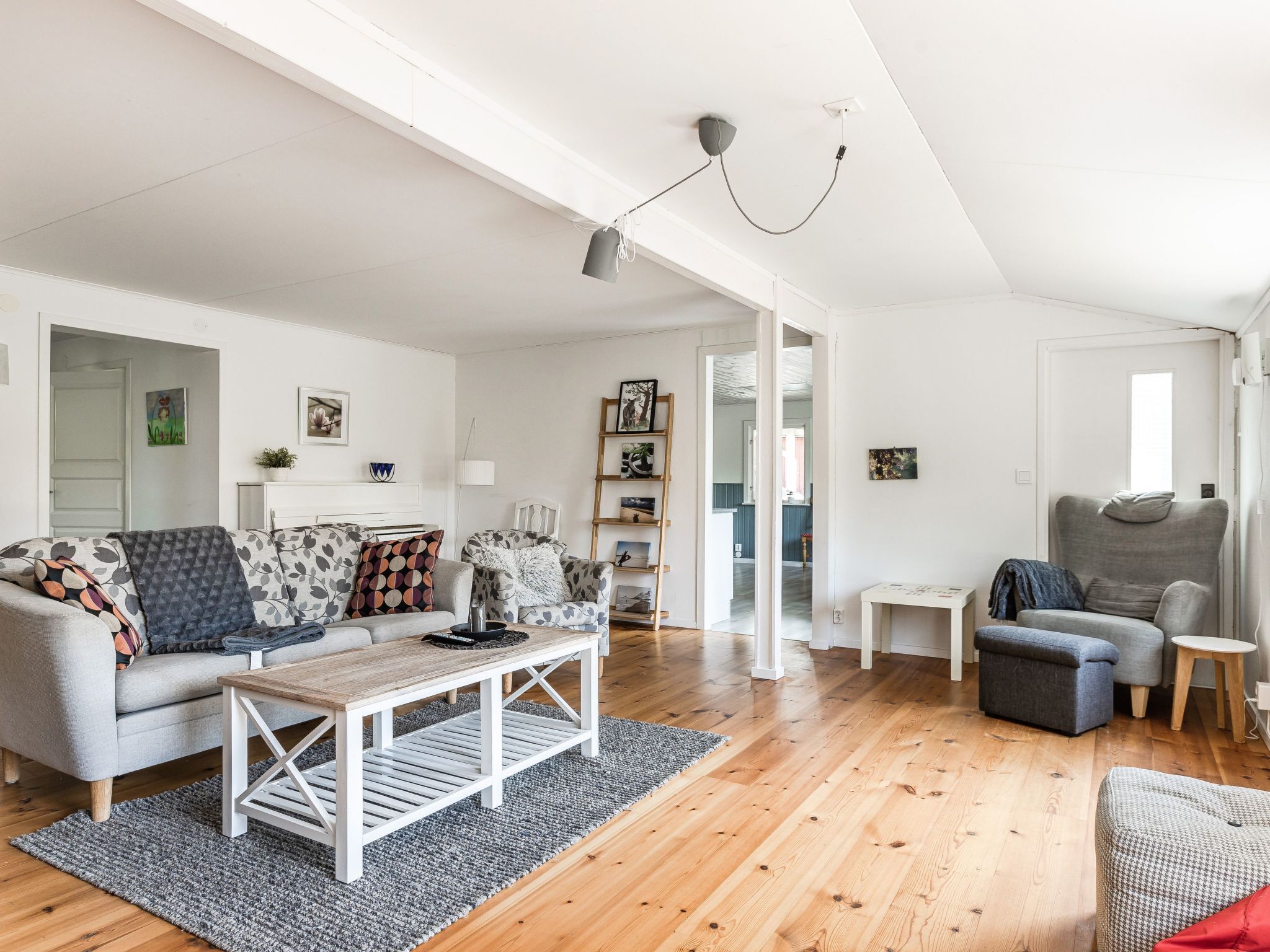 En rummelig stue med sofa, sofabord og et lille sidebord. En lænestol står ved vinduet, og der kan ses en reol i baggrunden.