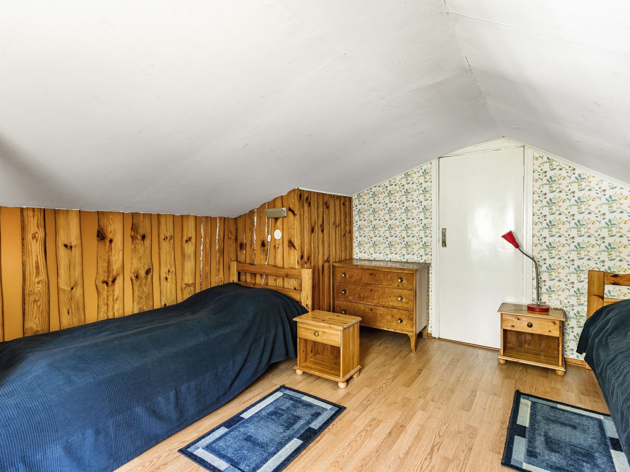 A small, cozy room with a sloped ceiling, wooden walls, two beds, a dresser, and a nightstand with a lamp.