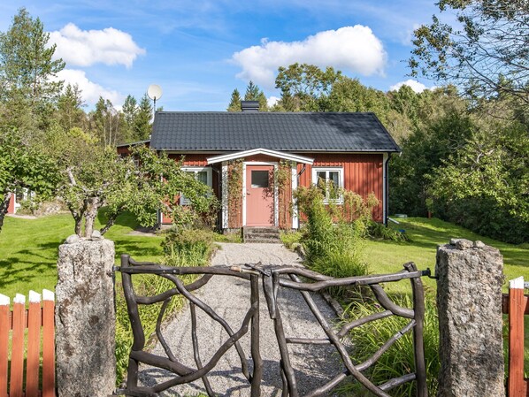 A red wooden house with a black roof, surrounded by greenery and a gravel path leading to the entrance.