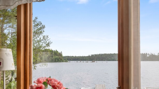 Une vue depuis une fenêtre donnant sur un lac, avec un quai et un bateau, le tout entouré d’arbres.