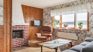 Un salon chaleureux avec une cheminée en briques, un fauteuil en cuir, un canapé, une table recouverte d’une nappe blanche et une fenêtre donnant sur l’extérieur.