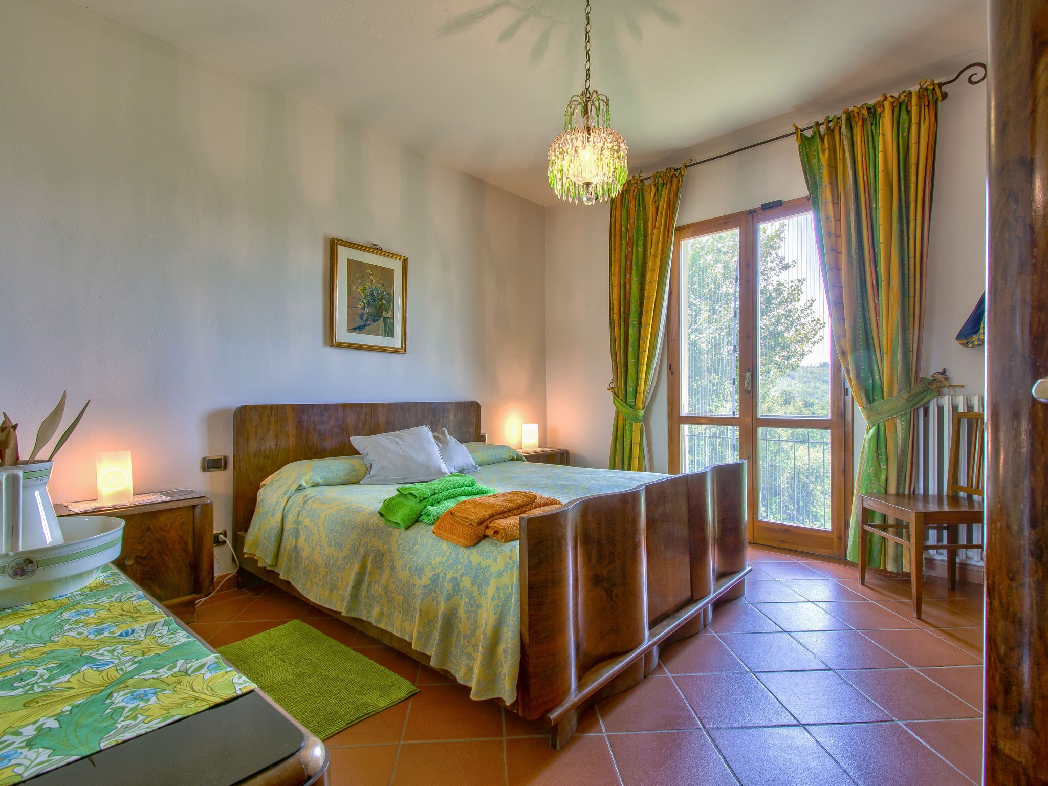 A bedroom with a wooden bed, a bedside table, a nightstand, a green rug, a window with curtains, and a hanging light fixture.