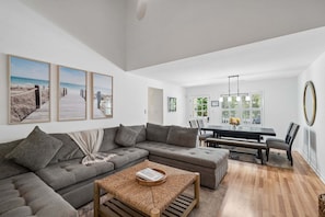 A modern living room with a sectional sofa, a dining table with chairs, and a large window with blinds.