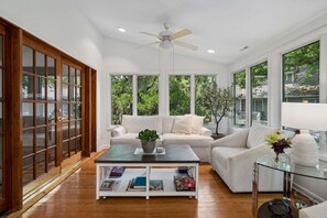 Un salon spacieux avec une grande fenêtre, un ventilateur de plafond et un canapé blanc.