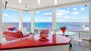 A modern room with a large window overlooking the ocean, a red bar area, and a seating arrangement with red chairs.