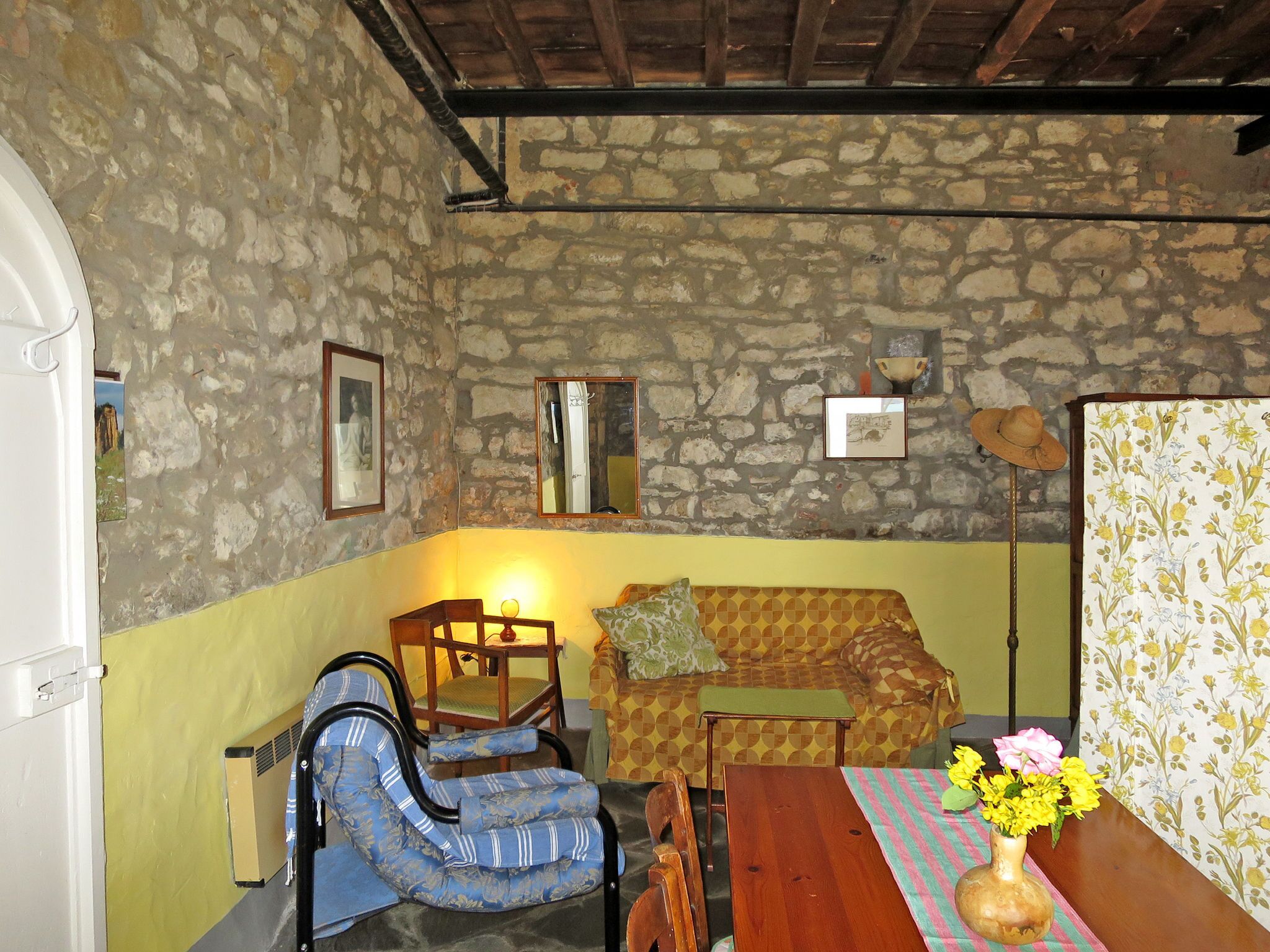 Una habitación acogedora con pared de piedra, techo de madera, un sofá estampado, un sillón azul, una mesa de madera y un jarrón con flores.