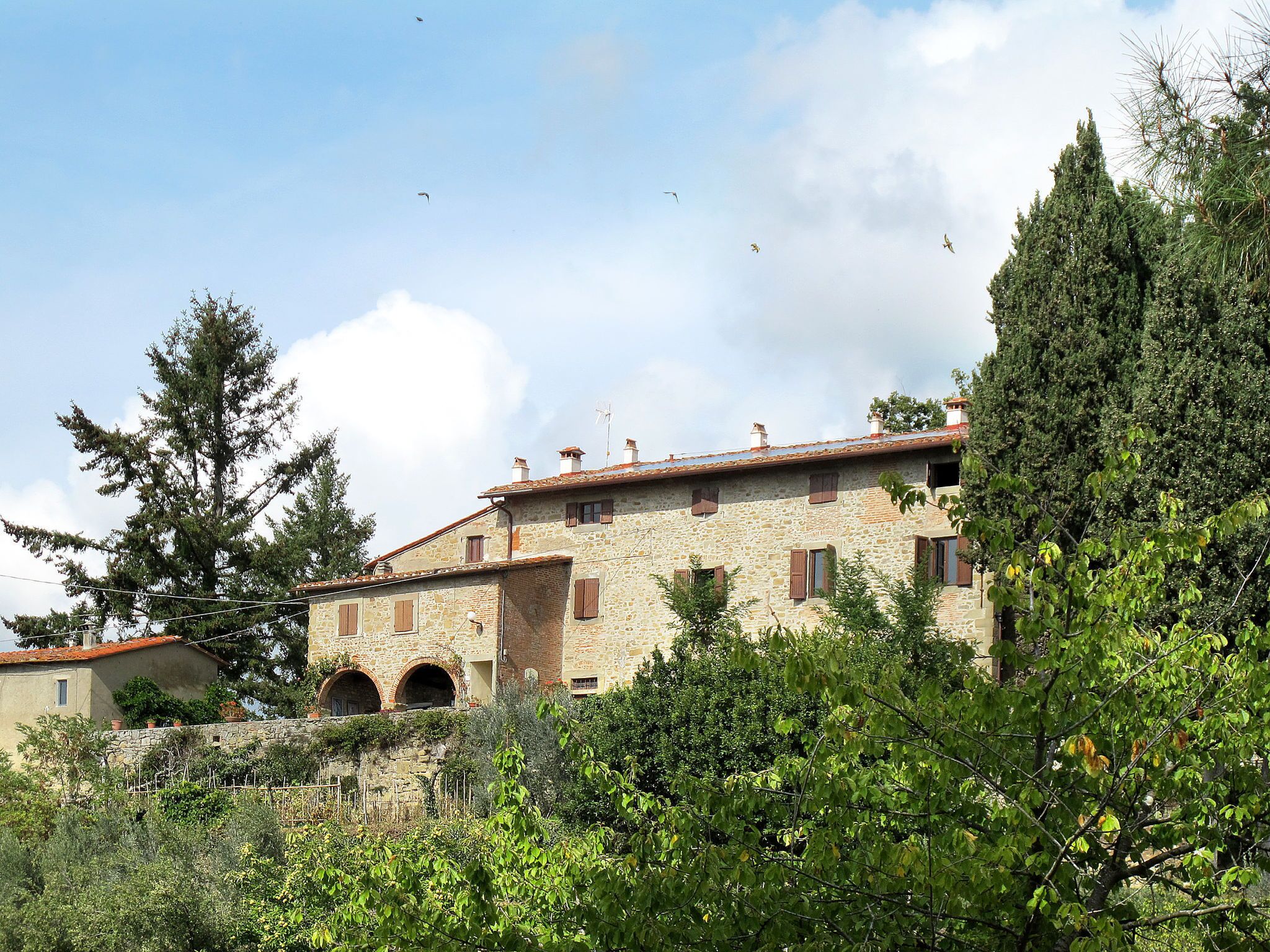 A stone building with arched windows and a red-tiled roof, surrounded by greenery and trees.