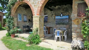 A rustic stone building with two arches, a small outdoor seating area with white plastic chairs, and a potted plant.