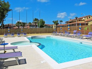 A swimming pool with sun loungers and umbrellas, surrounded by a fence and trees. - Riviera 3 by Interhome (Cecina)