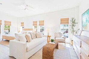 A spacious living room with a large sofa, a coffee table, and a TV. There are windows with blinds, a ceiling fan, and a plant.