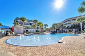 A swimming pool with fountains, surrounded by lounge chairs and palm trees, with buildings in the background.