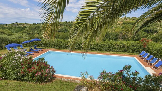 A swimming pool surrounded by blue lounge chairs and umbrellas, with lush greenery and a clear sky in the background. - Carbonaia (PAA133) by Interhome (Palaia)