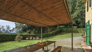 A wooden picnic table with benches under a thatched roof, surrounded by a grassy area with a view of a hill and trees. - Favilli (PAA130) by Interhome (Palaia)