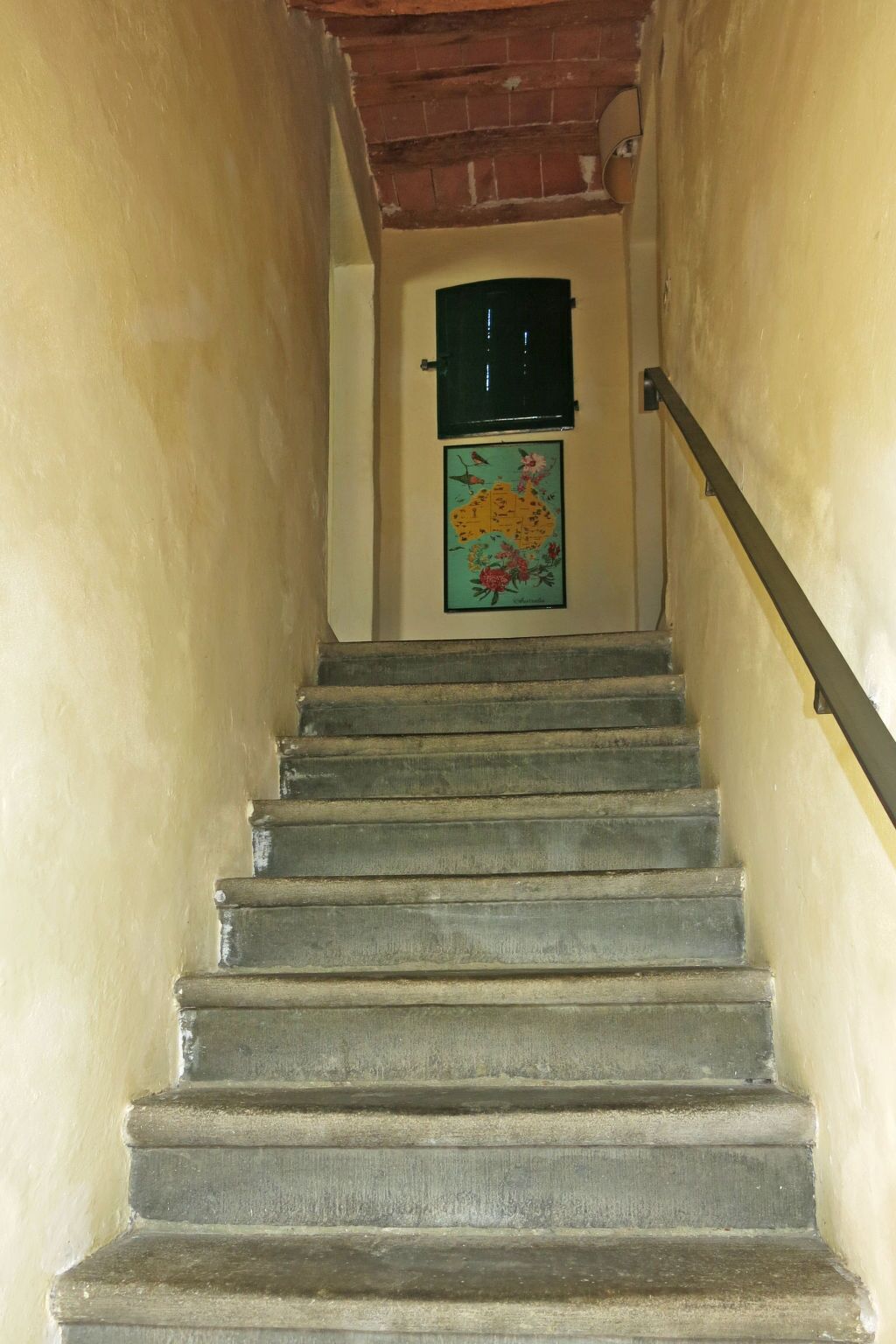 A staircase with a handrail, leading to a door with a green shutter and a floral artwork below.