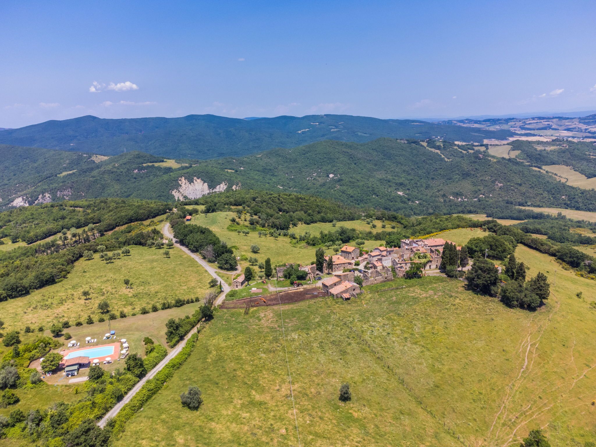 An aerial view of a rural village with a central cluster of stone buildings, surrounded by green fields and hills.