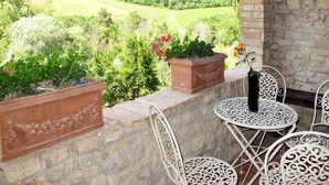 Een terras met stenen muren, witte metalen stoelen en terracotta plantenbakken met bloemen.