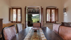 Een eetruimte met een lange tafel, stoelen en uitzicht op een groen landschap door open ramen.