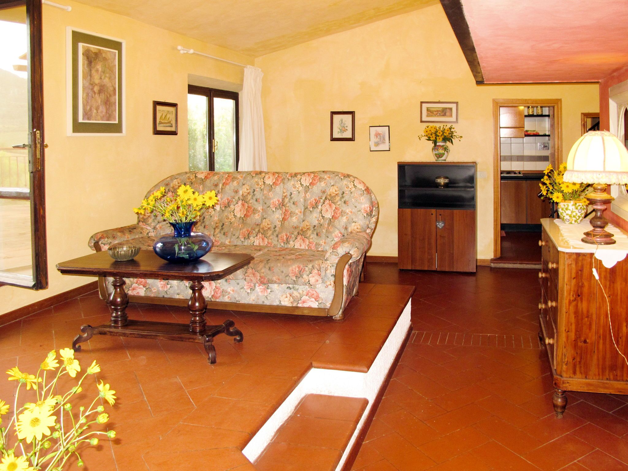 A living room with a floral sofa, a wooden table with a vase of flowers, and a kitchen area in the background.