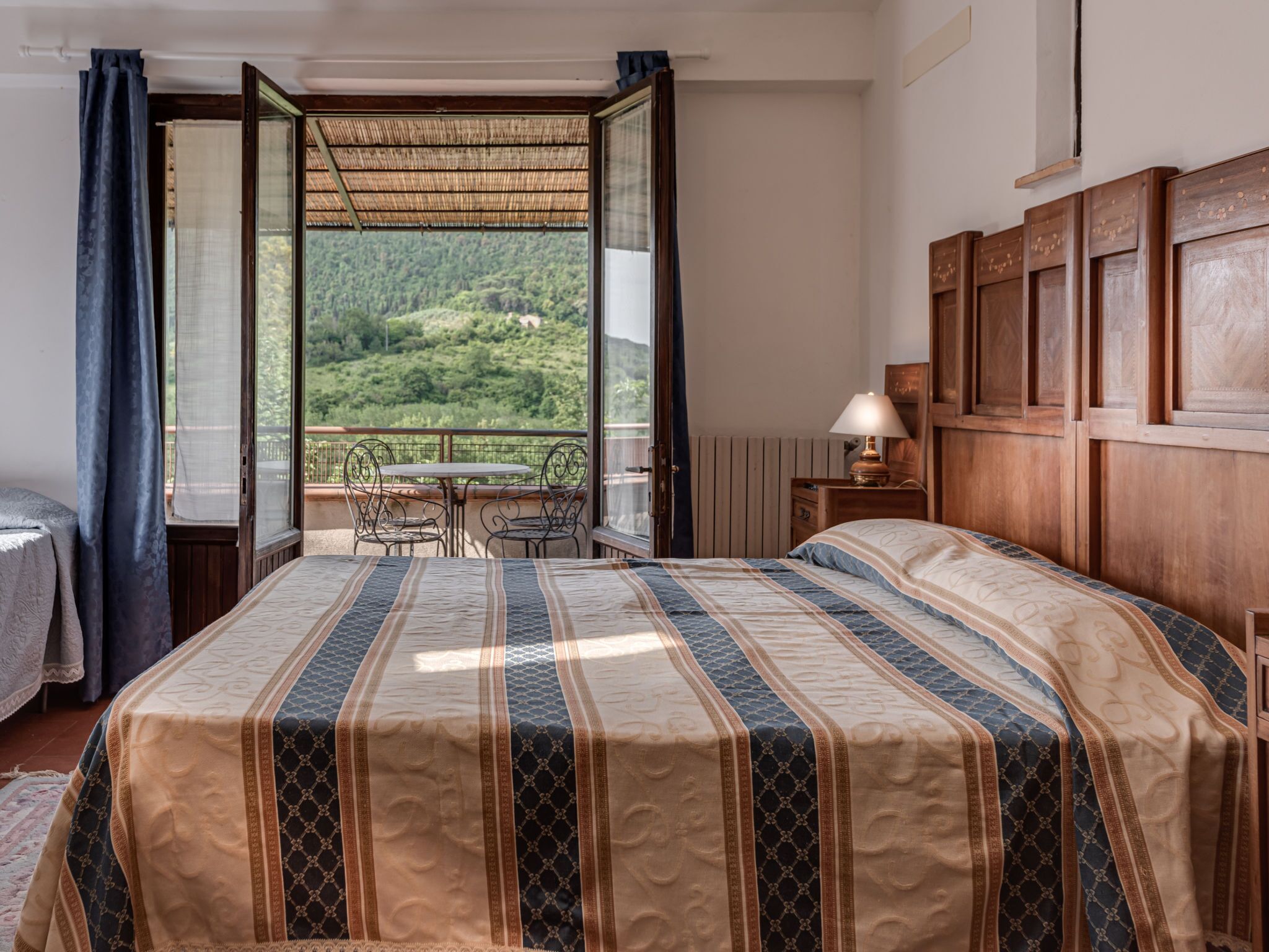 A bedroom with a large bed, a wooden headboard, a small table with chairs, and a balcony with a view of a green landscape.