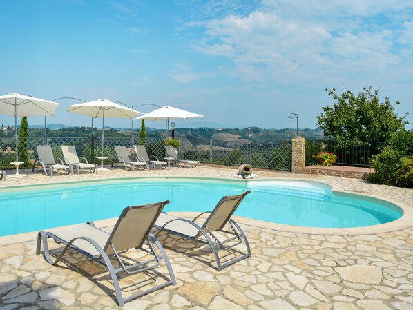 Zona de piscina con sillones de descanso y sombrillas, rodeada de baldosas de piedra y vistas al campo.