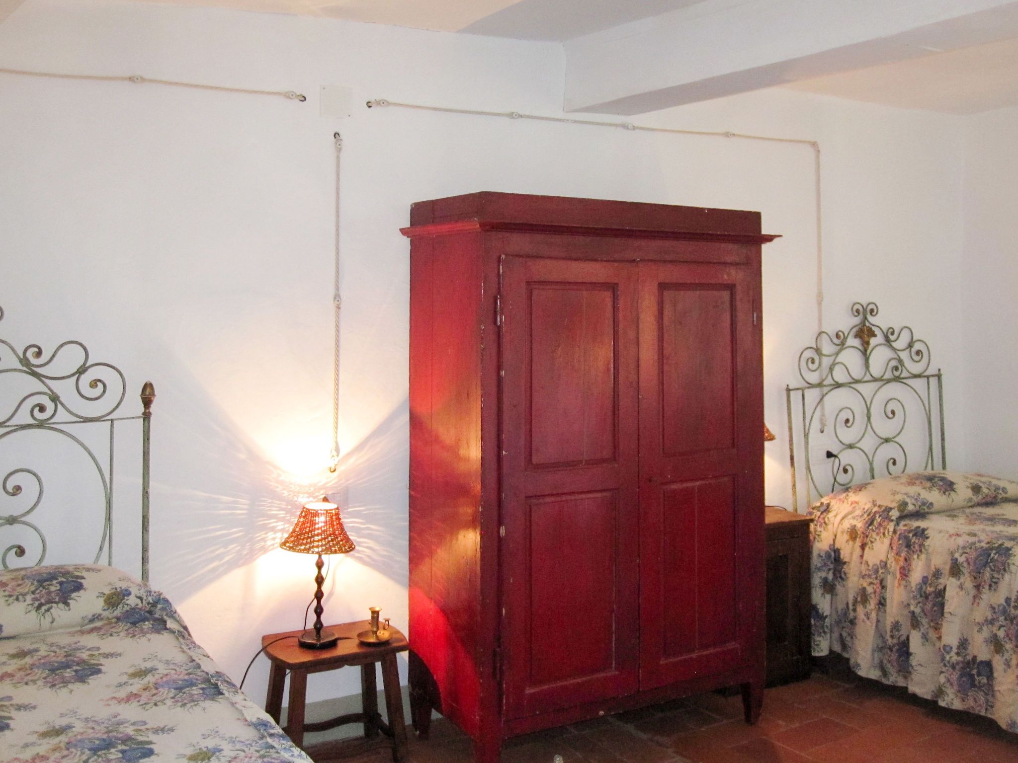 A bedroom with a wooden wardrobe, a bed with floral bedding, a bedside table with a lamp, and a metal headboard.