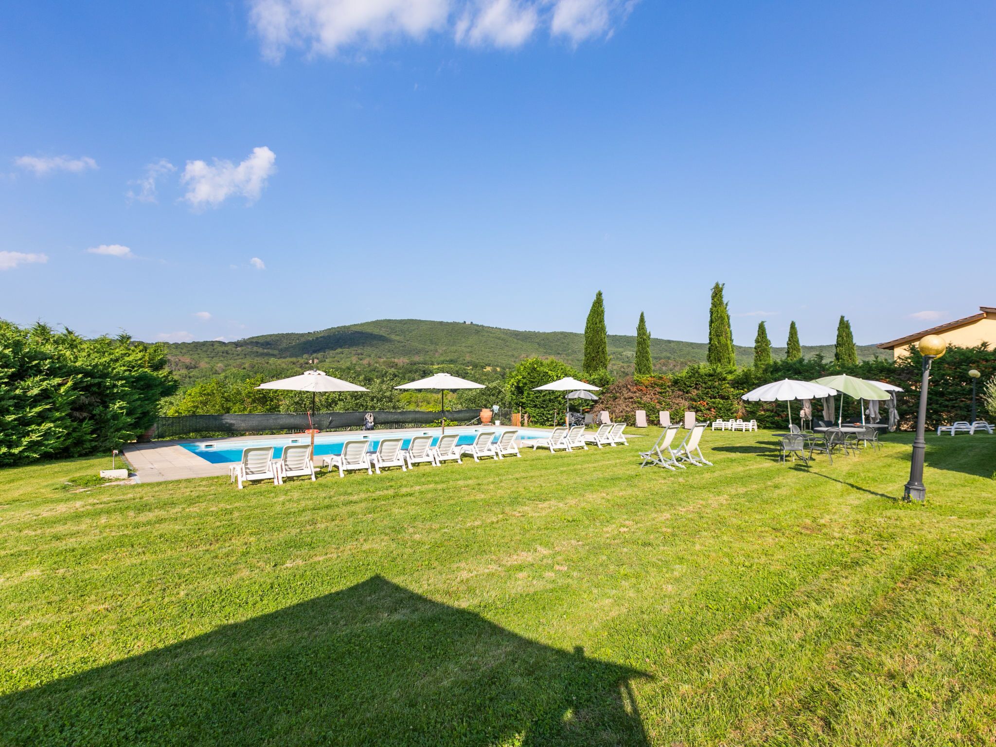 A pool area with lounge chairs and umbrellas, surrounded by grass and trees, with a mountain in the background.