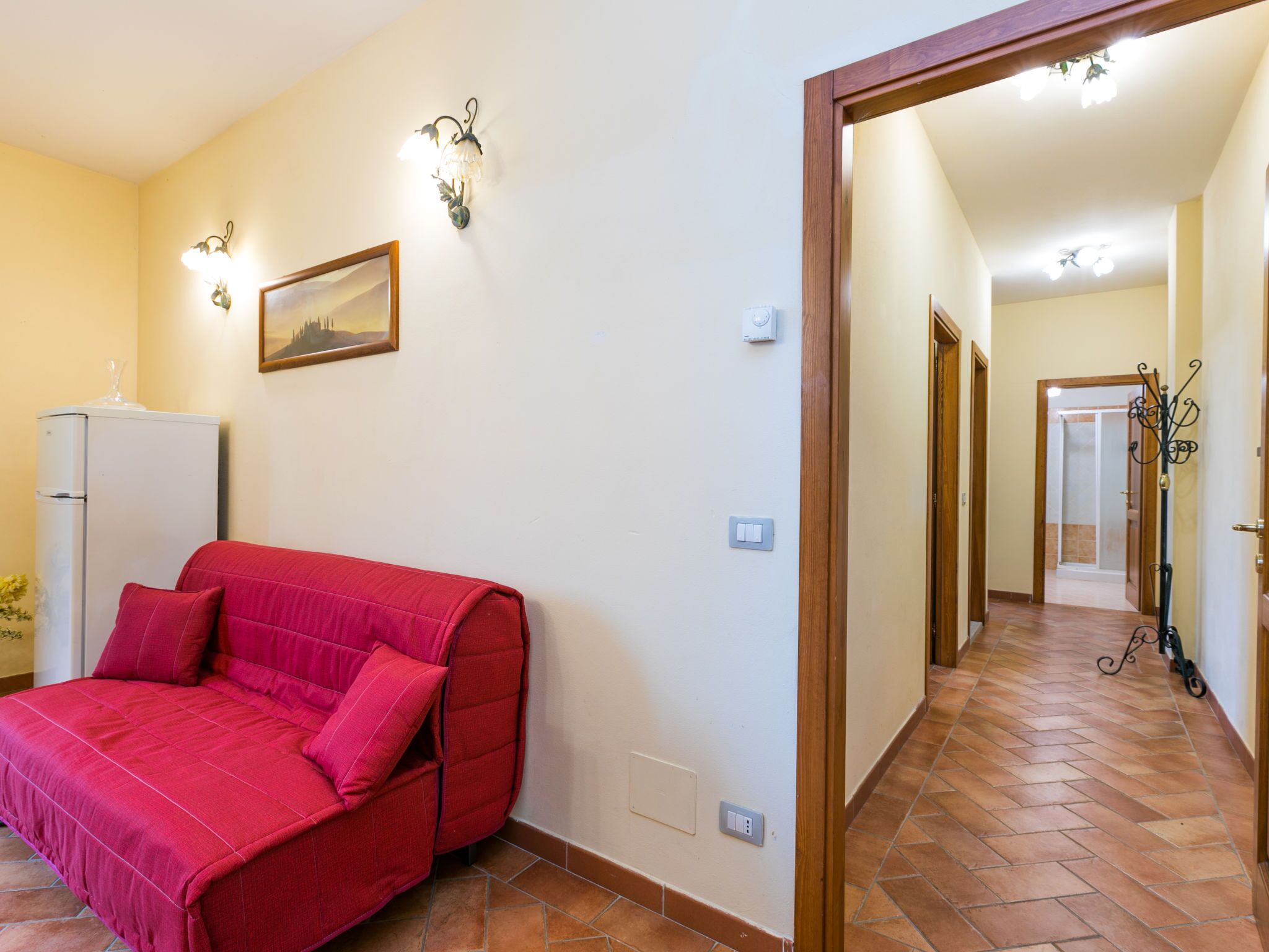A room with a red sofa, a white refrigerator, a wall-mounted light fixture, and a door leading to another room.