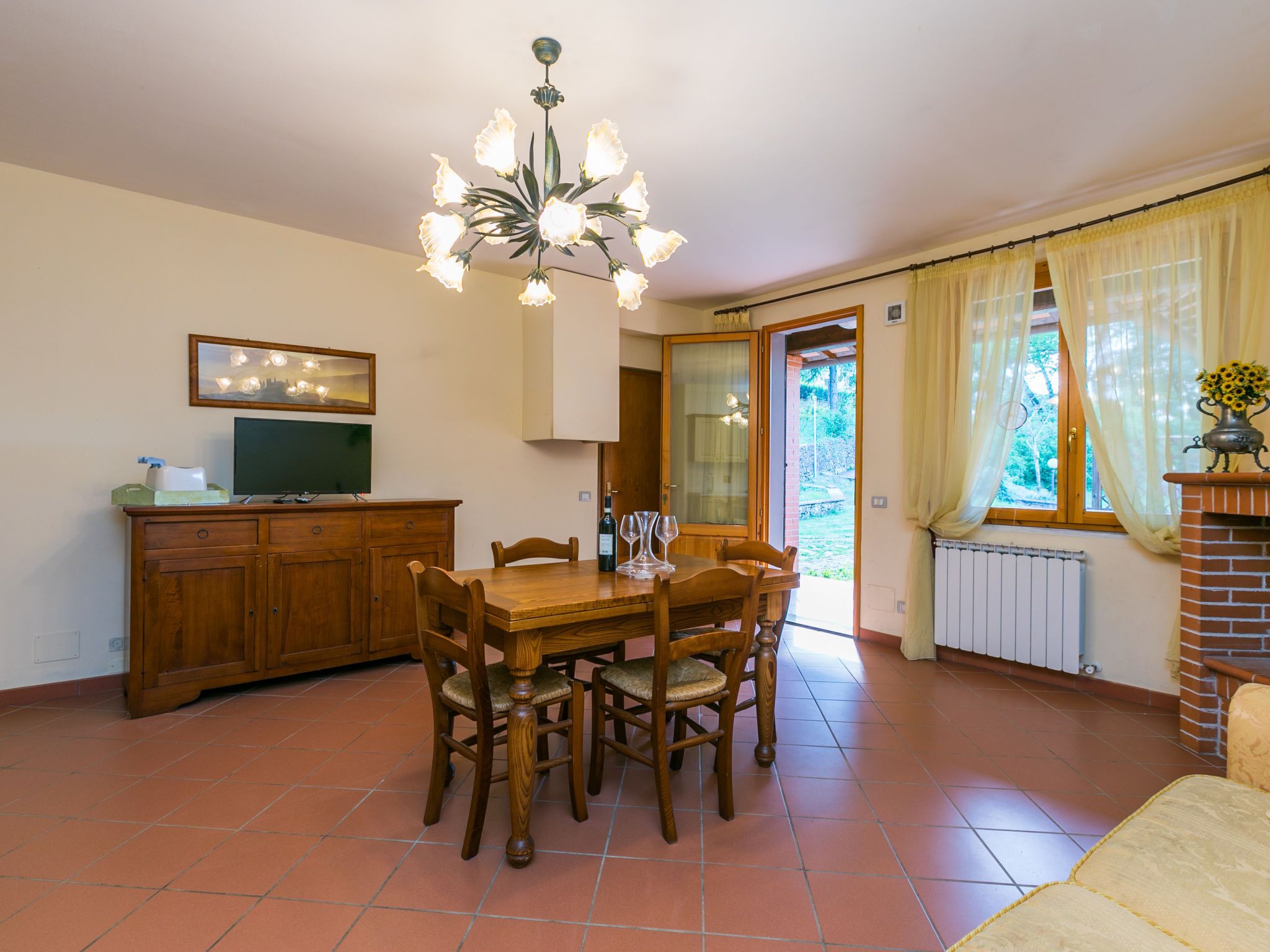 Un coin repas avec une table et des chaises en bois, un lustre, un téléviseur posé sur un meuble et une vue sur l’extérieur par une porte.