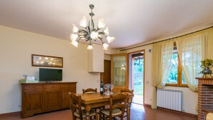 Zona de menjador amb taula i cadires de fusta, un lampadari, un televisor sobre un moble i vista a l'exterior a través d'una porta.