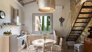 Une cuisine avec une table de salle à manger ronde, des chaises en bois, un luminaire suspendu, une fenêtre donnant sur des arbres et un escalier en pierre.