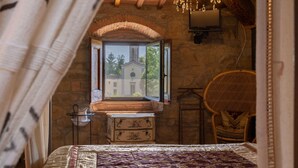 Un dormitorio con pared de piedra, techo de madera, cama con colcha estampada, cómoda, silla de mimbre y ventana con vista a un edificio.