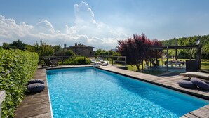 Una piscina rectangular con cubierta de madera, rodeada de vegetación y una casa al fondo.